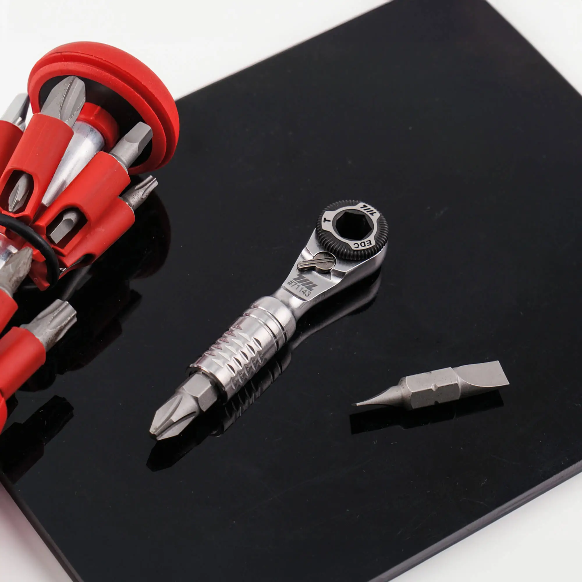 Mini ratchet wrench on a black work surface next to a red-handled screwdriver and spare bit